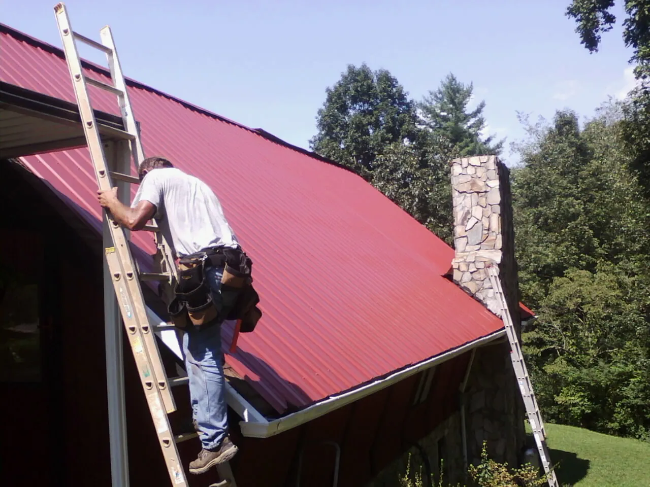 Roofer on a ladder beside a red metal roof and stone chimney — Soffit & Fascia Repair by Roofing Brothers in North Andover